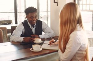 Black Man and Red-Haired woman seated with cups while making notes